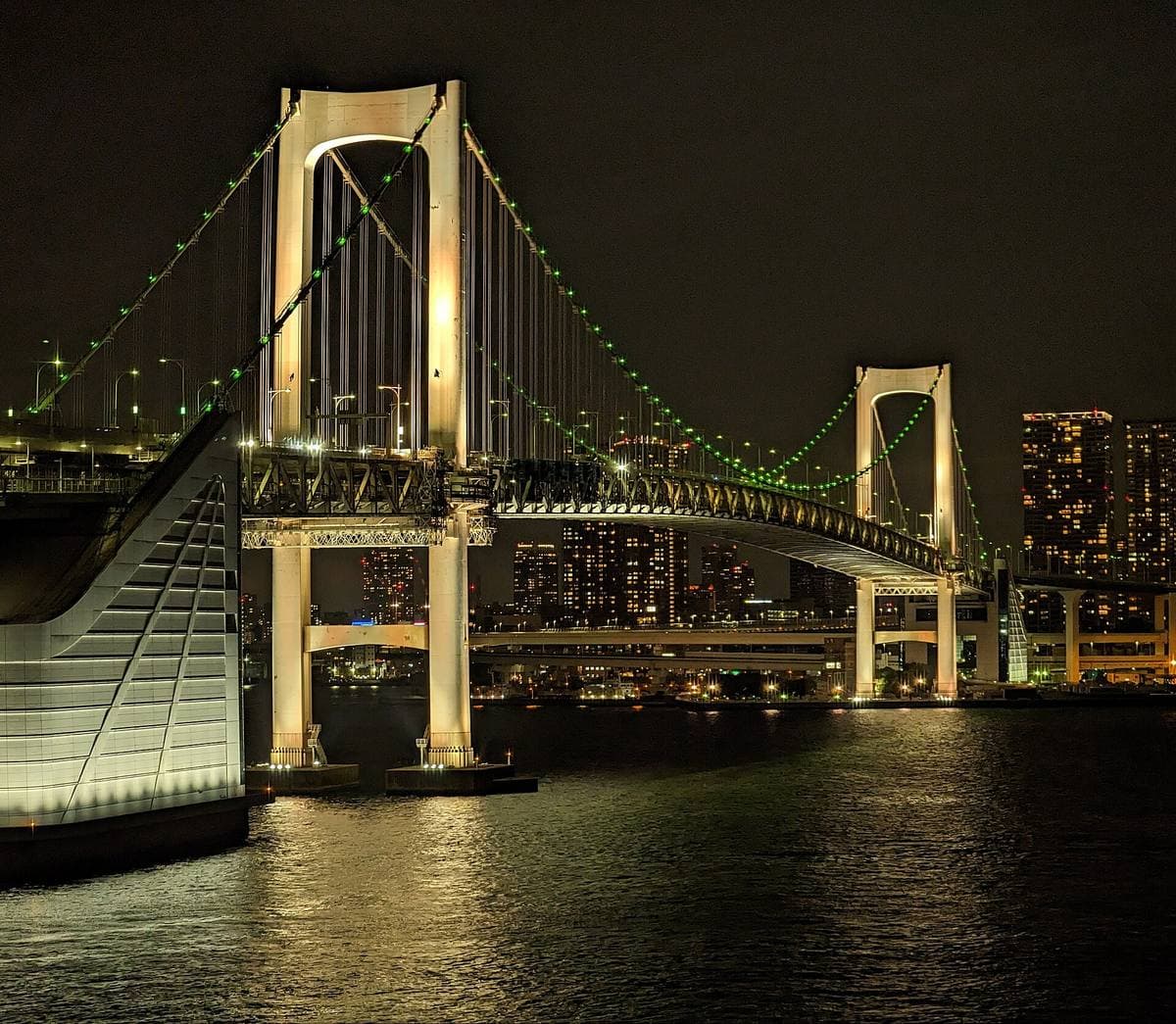 A lit suspension bridge over dark water with city lights behind