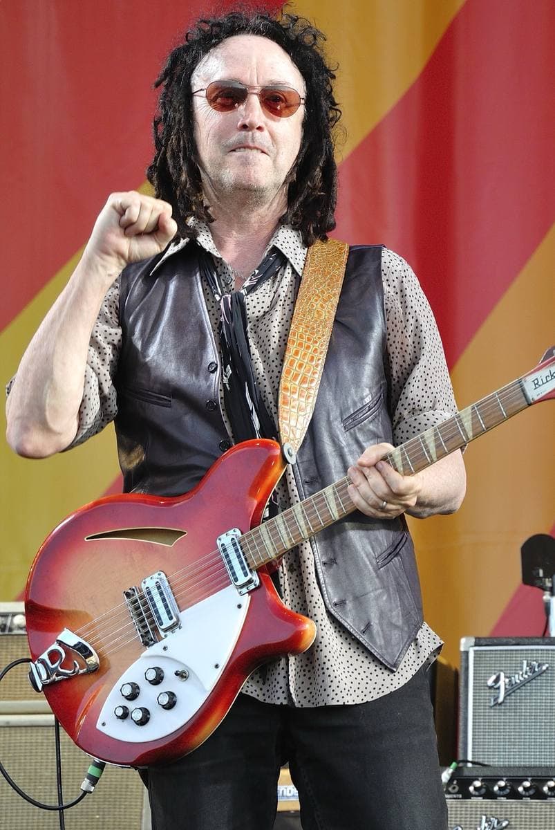 Mike Campbell in sunglasses and a leather waistcoat raising a fist, a red Rickenbacker guitar strapped on