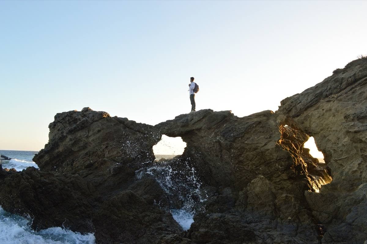 A figure standing on dark coastal rocks above a sea arch with waves below