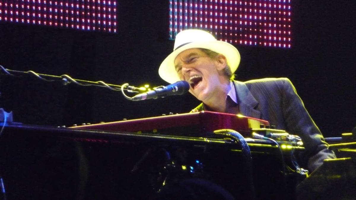 Benmont Tench in a white hat singing at a keyboard under stage lights