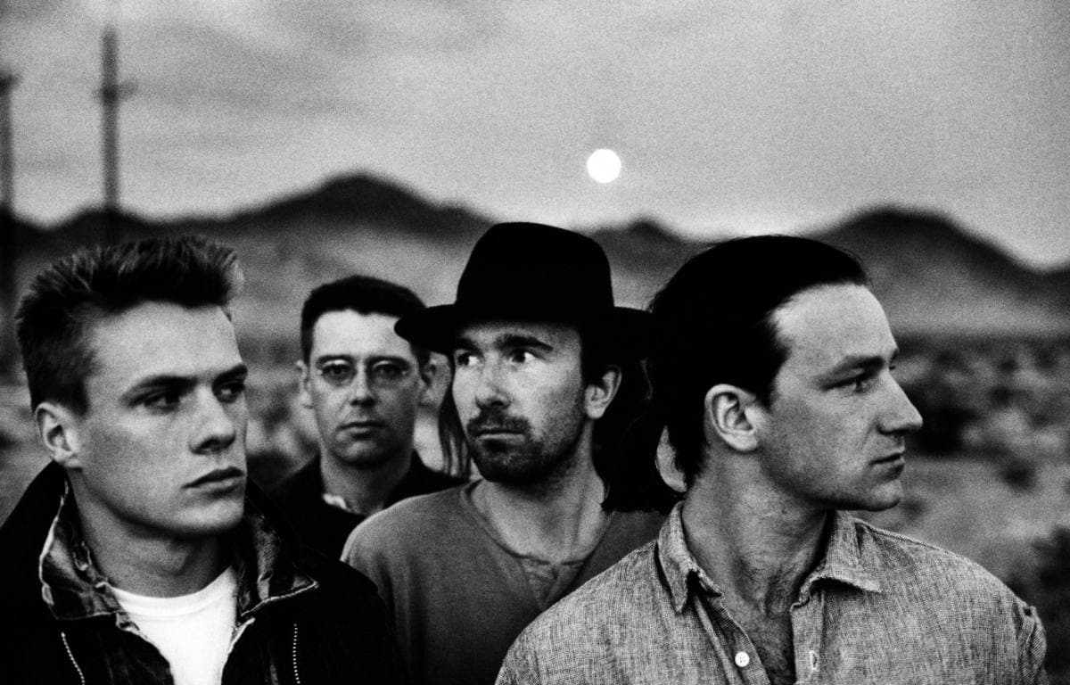 The four members of U2 in dark coats and hats against a desert ridge, in black and white