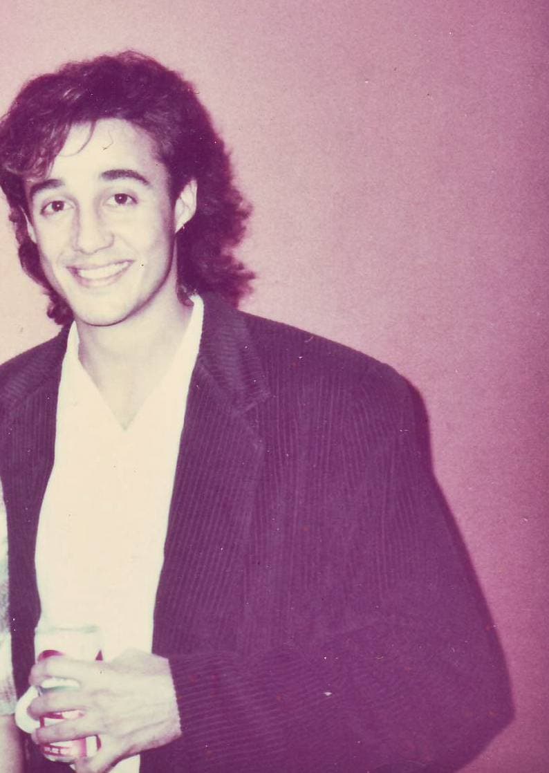 A smiling Andrew Ridgeley with long dark hair, a white shirt under a dark corduroy jacket, holding a drink, against a pink wall
