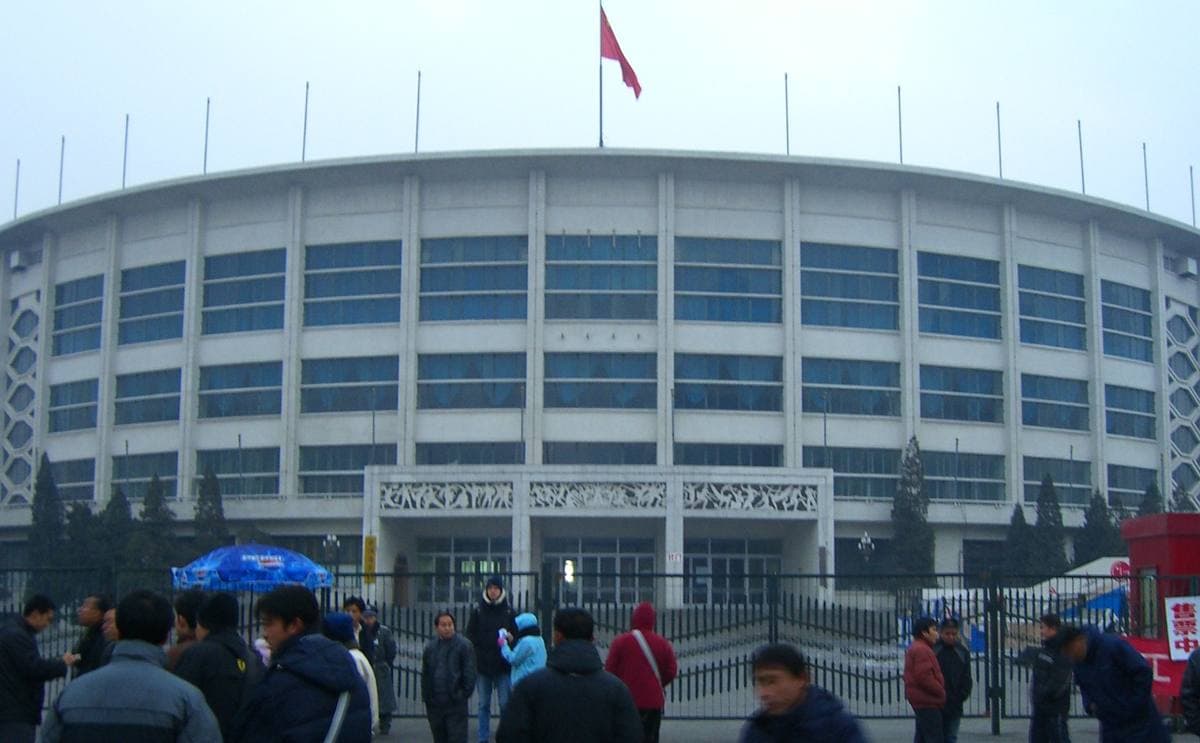A round, many-windowed arena with a red flag on its roof and people walking in front