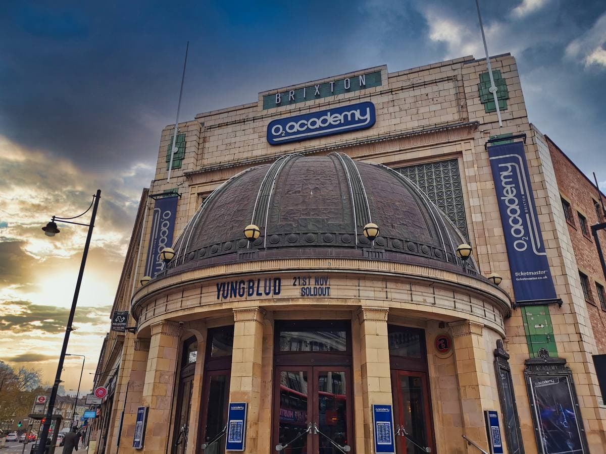 The domed corner entrance of Brixton Academy under a sunset sky, its name in lights