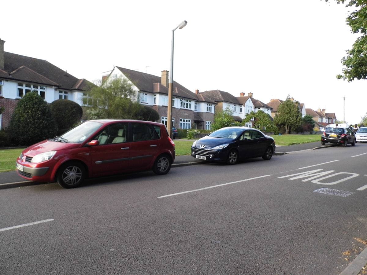A suburban road of 1930s semi-detached houses with cars parked along the kerb