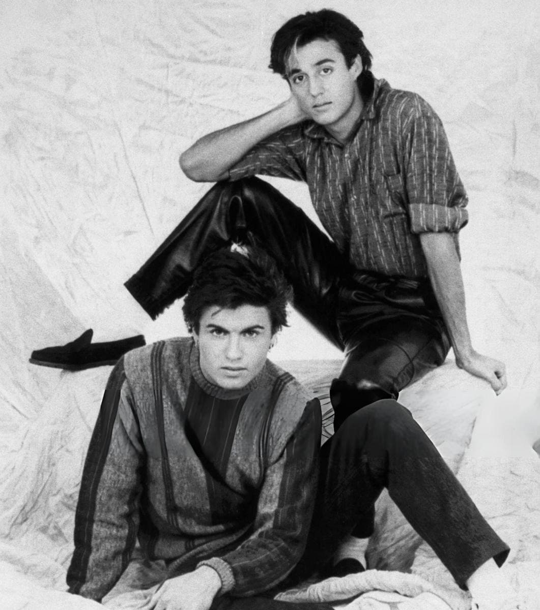 Two young men posed on a crumpled pale backdrop in black and white, Andrew Ridgeley seated high behind with his head on his hand, George Michael in front in a striped jumper