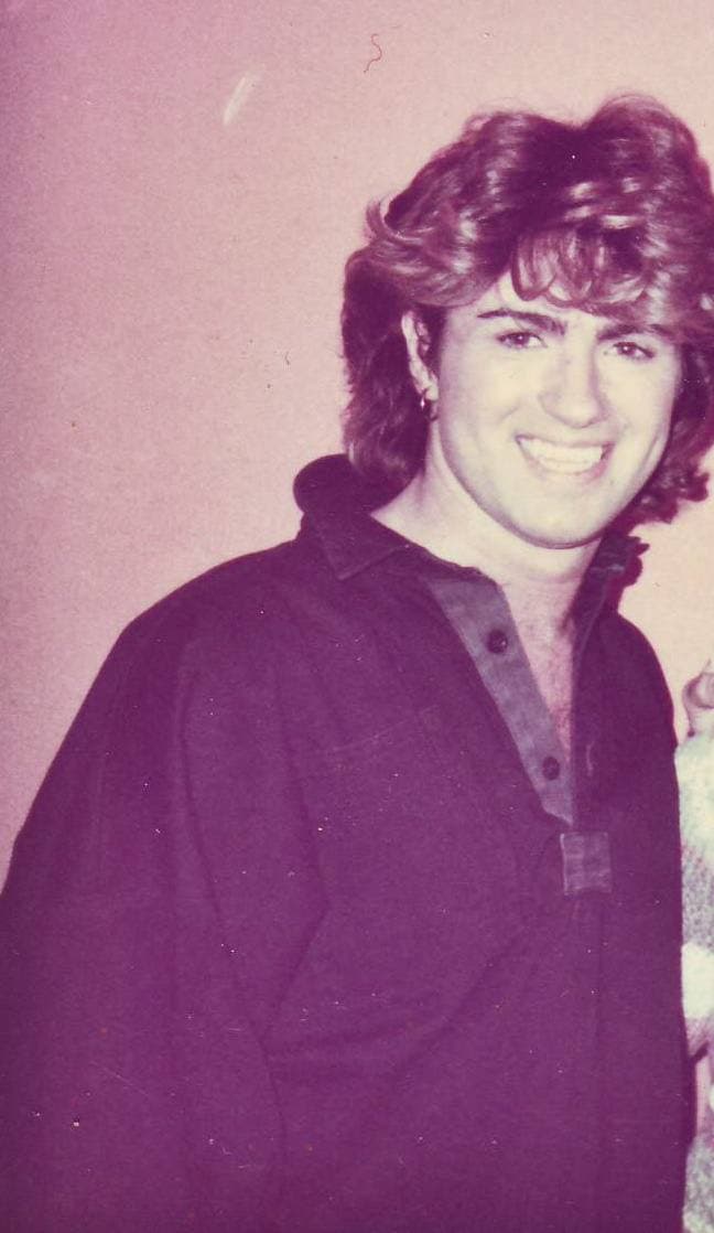 A smiling George Michael with a big, feathered 1980s mane, in a dark shirt open at the collar, against a pink wall
