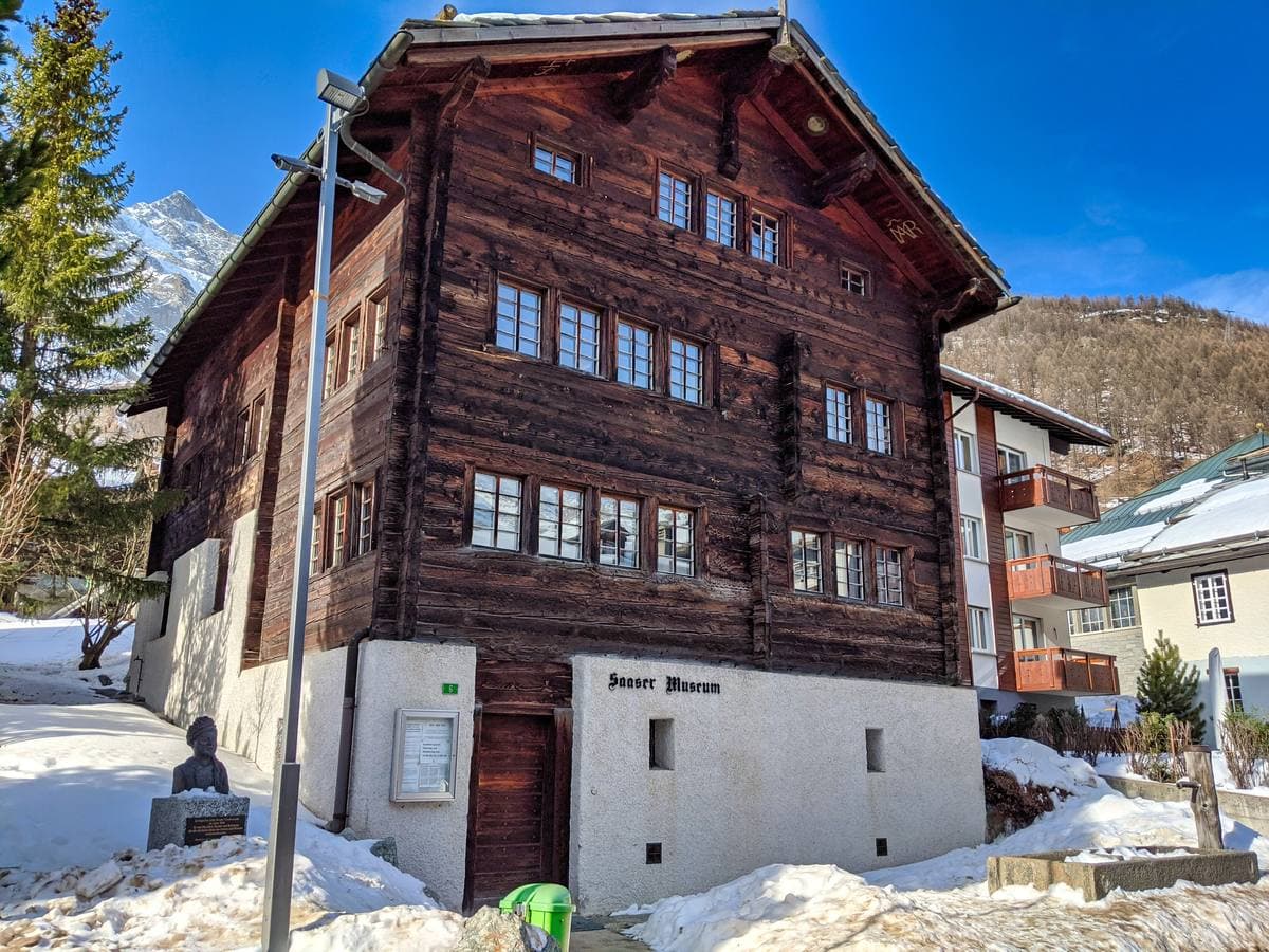 A tall dark-timbered house on a white stone base, snow on the ground, in sunshine