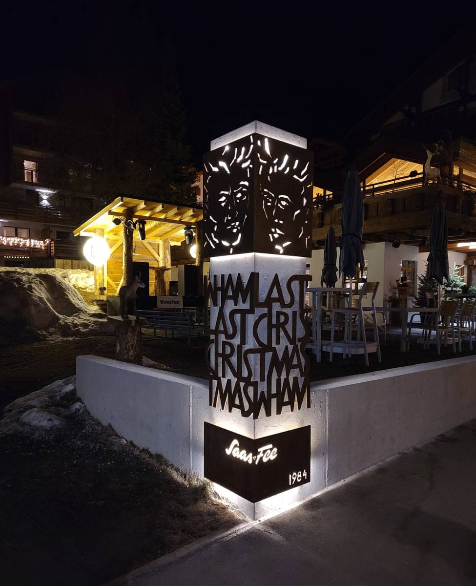 A tall lit pillar at night cut with two faces and the words of the song title, beside a hotel terrace