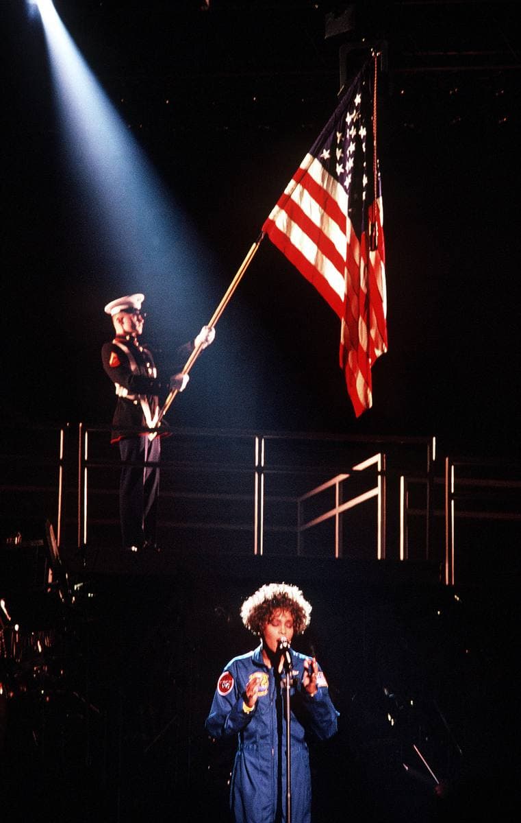 Whitney Houston in a blue flight suit sings on a stage below a color guard holding the American flag