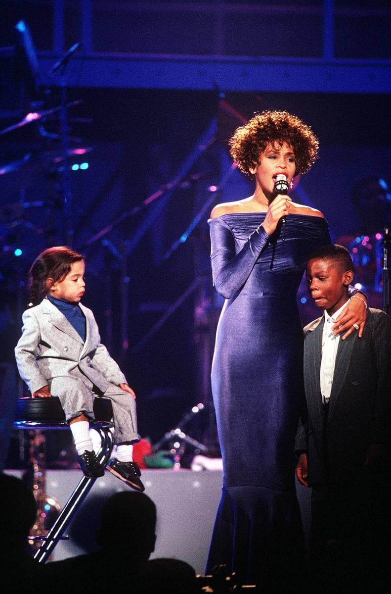 Whitney Houston in a long blue gown singing with two children beside her on a lit stage