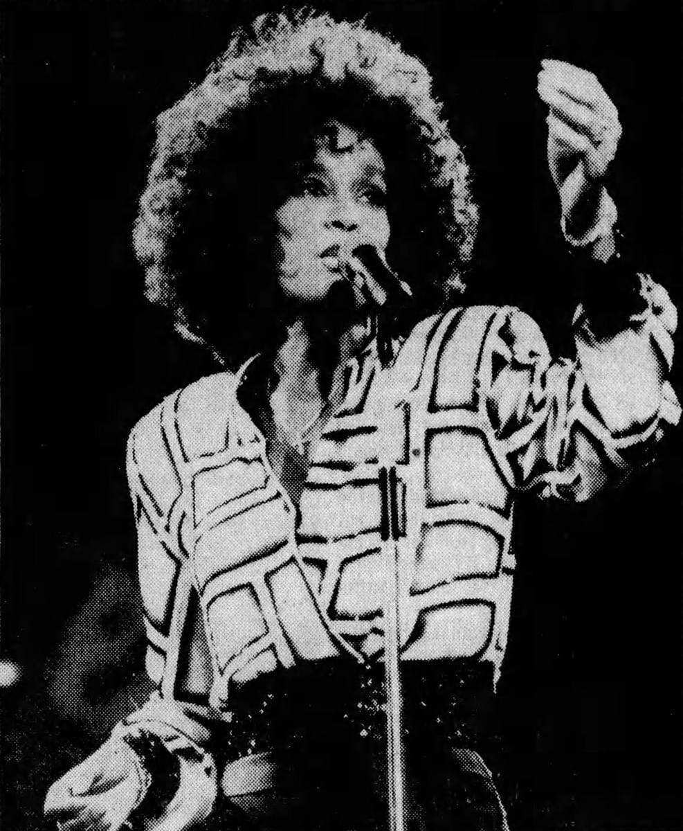 Whitney Houston singing into a stand microphone, one arm raised, in a black-and-white concert photograph