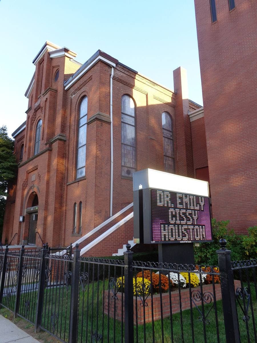 A red-brick church with tall arched windows behind an iron fence, a street sign reading Dr Emily Cissy Houston in front