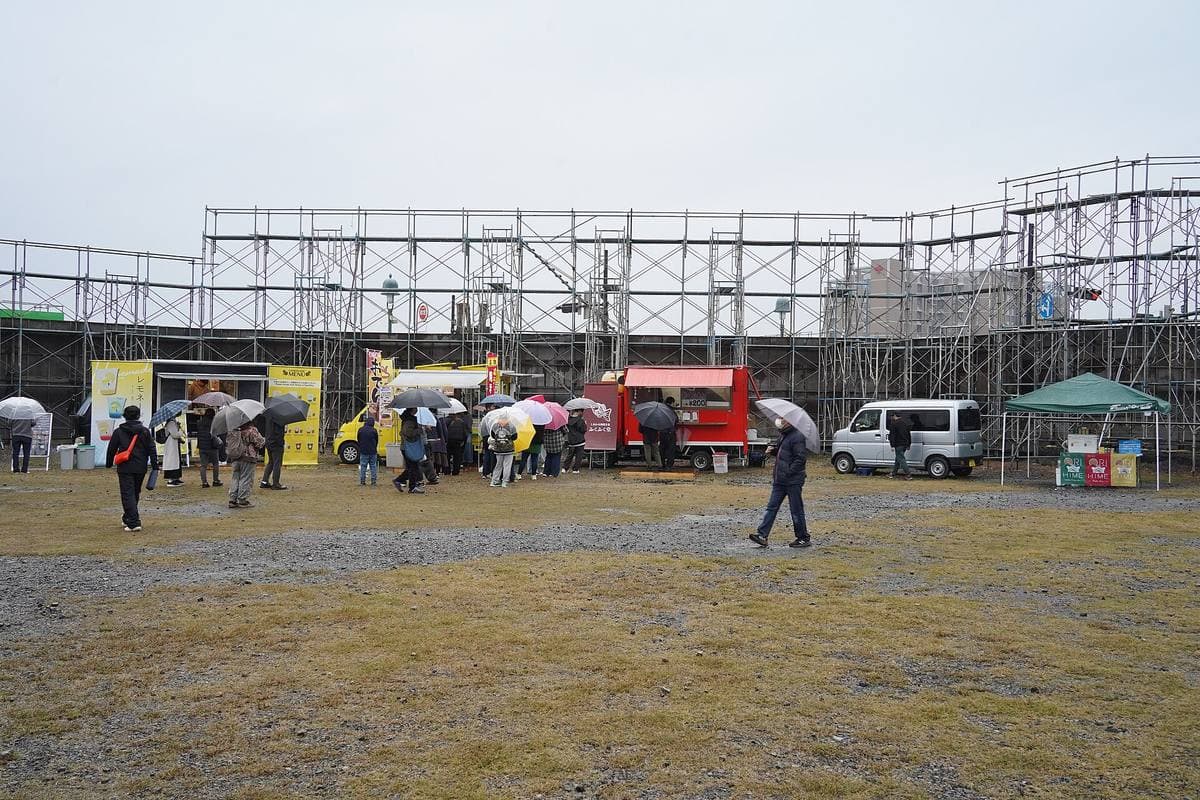 People with umbrellas among food vans on a wide grassy lot in front of tall scaffolding facades