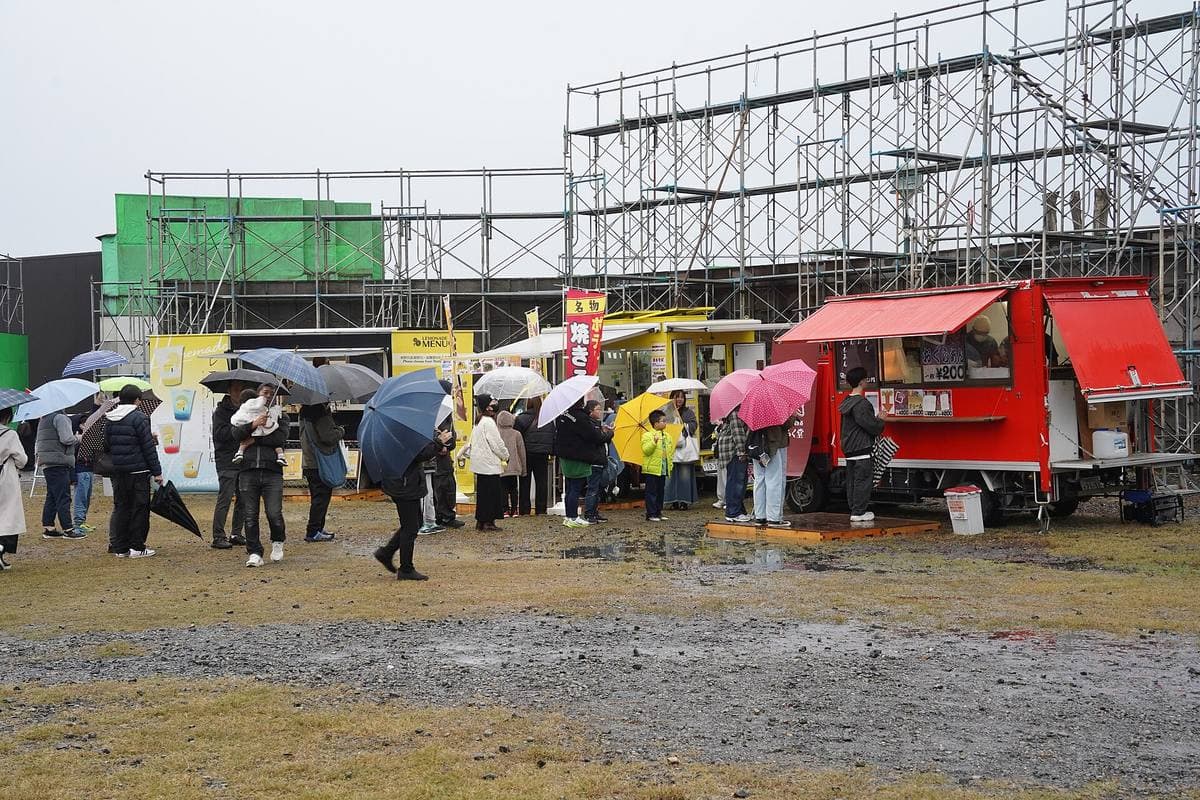 Visitors with umbrellas queue at red food vans in front of the set’s scaffolding