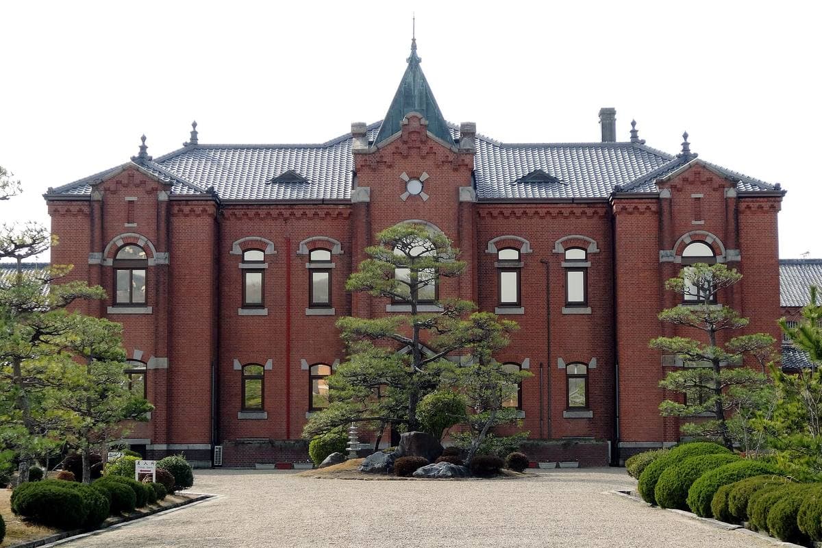 A red-brick Meiji building with a pointed central tower behind a clipped garden