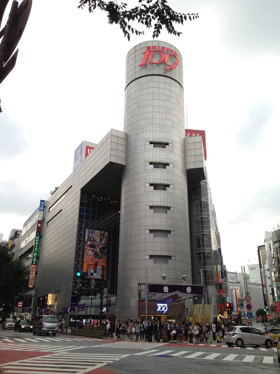 The silver cylindrical tower of Shibuya 109 with its red 109 sign, at a street corner