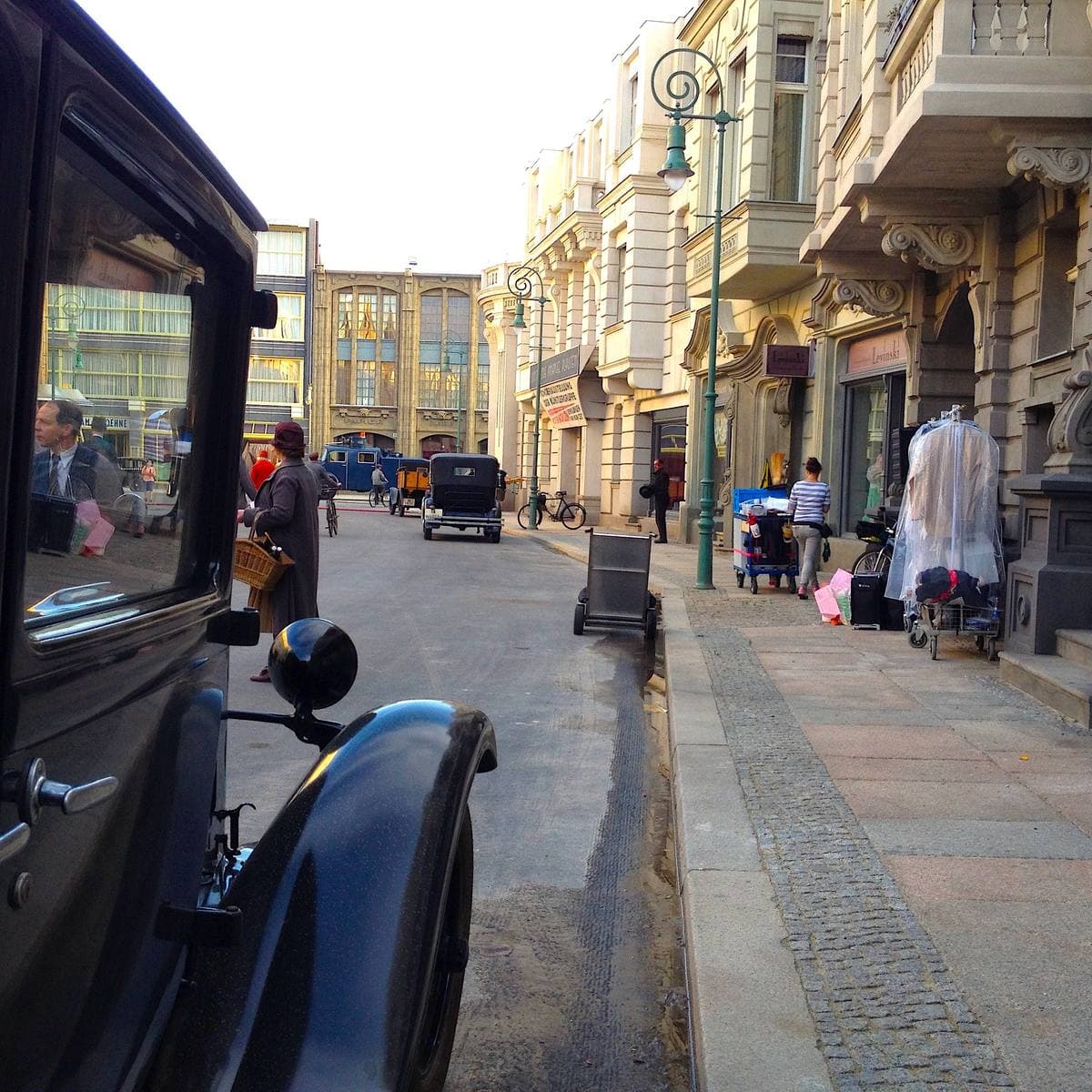 A black vintage car in the foreground of a set street with a clothes rail and people in period dress