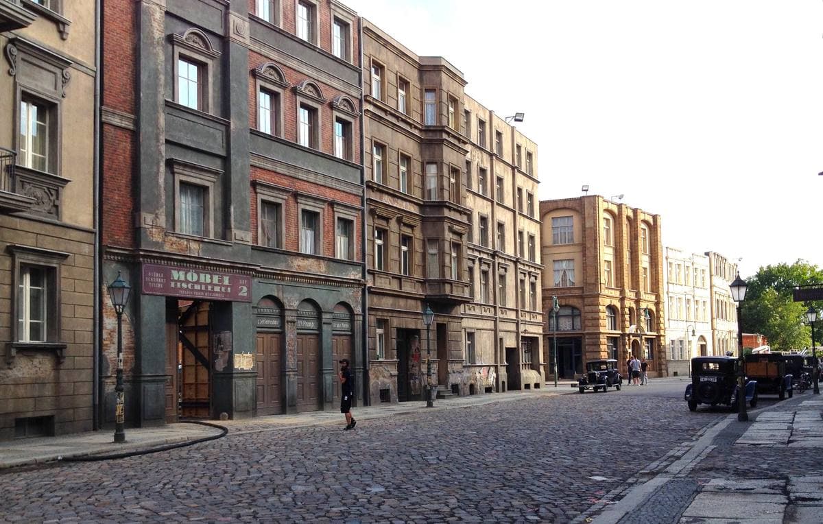 A cobbled street of 1920s tenement fronts with vintage cars parked at the kerb