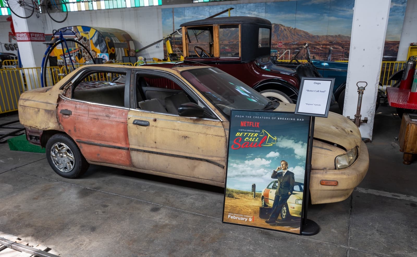 A battered pale yellow saloon car parked indoors beside a framed show poster