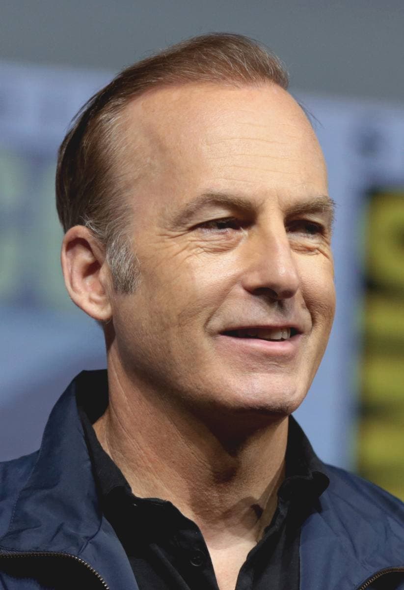 Bob Odenkirk speaking into a microphone at a panel table, in a dark jacket