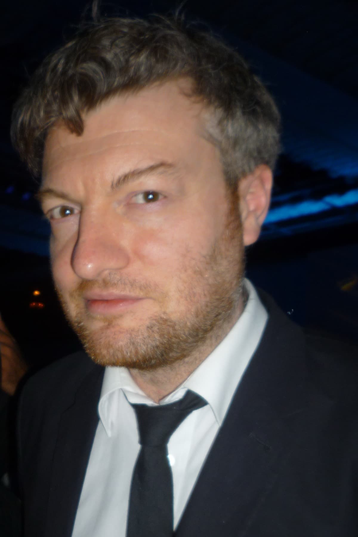 Charlie Brooker in a dark suit and tie against a dark background, looking at the camera