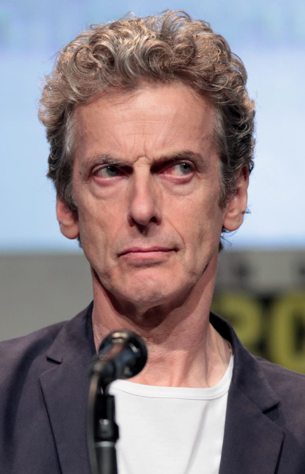 Peter Capaldi with grey curly hair, raising an eyebrow behind a microphone