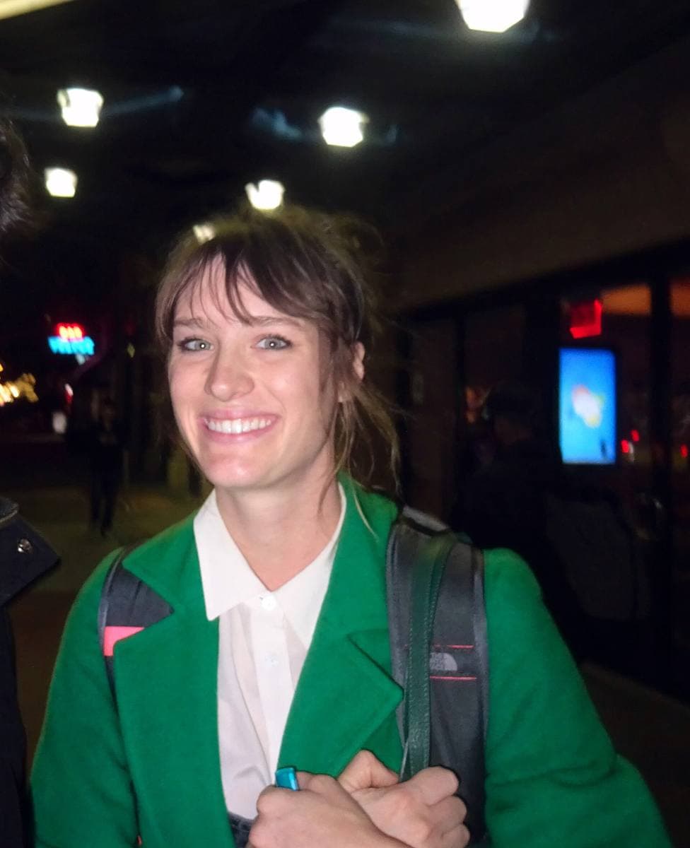 Mackenzie Davis smiling in a green jacket and white shirt on a city street at night