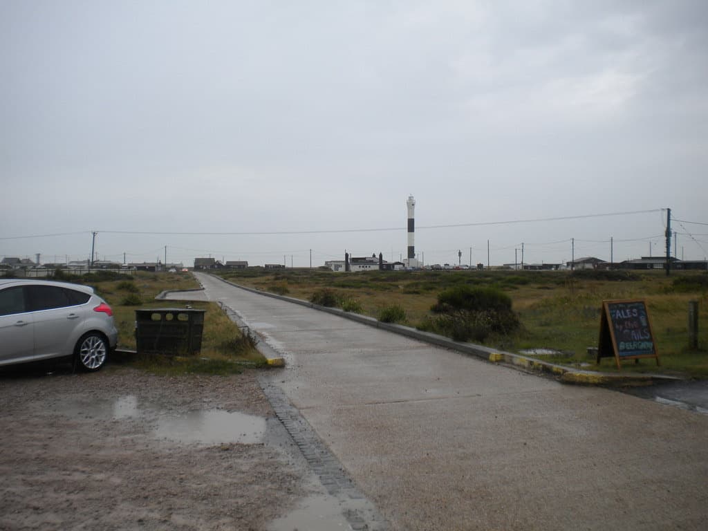 A wet concrete road running flat across shingle towards a lighthouse