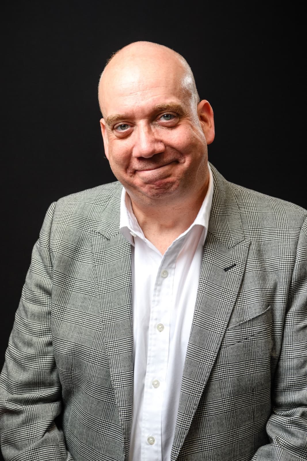 Paul Giamatti in a grey tweed jacket and white shirt, smiling against a dark background