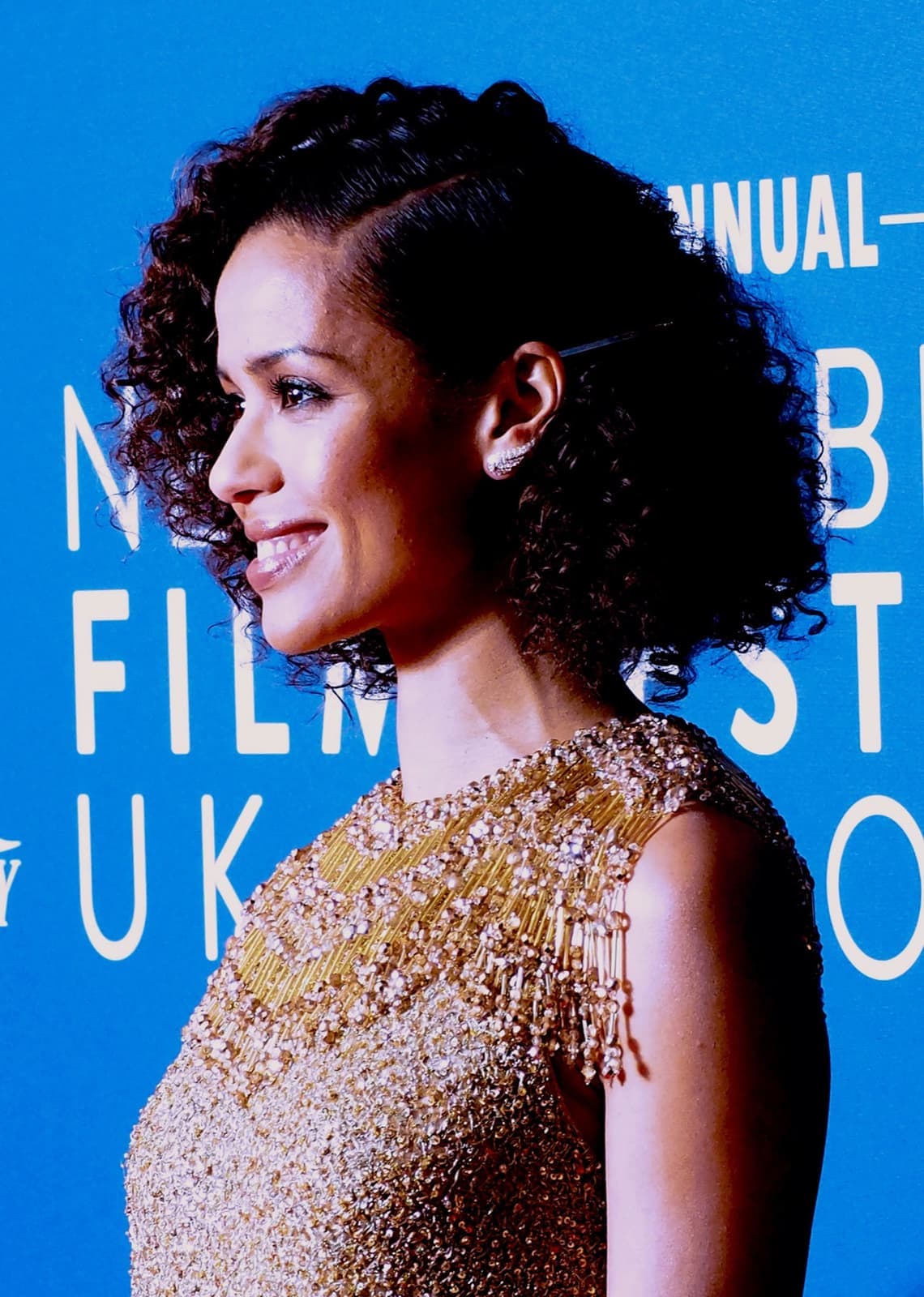 Gugu Mbatha-Raw in profile, smiling, in a gold beaded dress against a blue backdrop