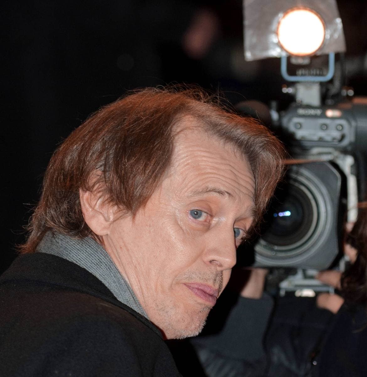 Steve Buscemi in a dark coat beside a television camera