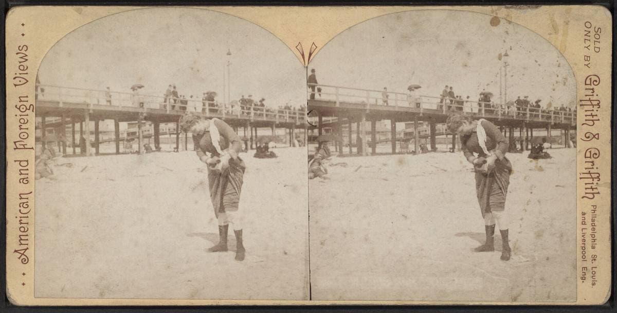A cream stereograph card with two arched photographs of a bather on the sand below a raised boardwalk