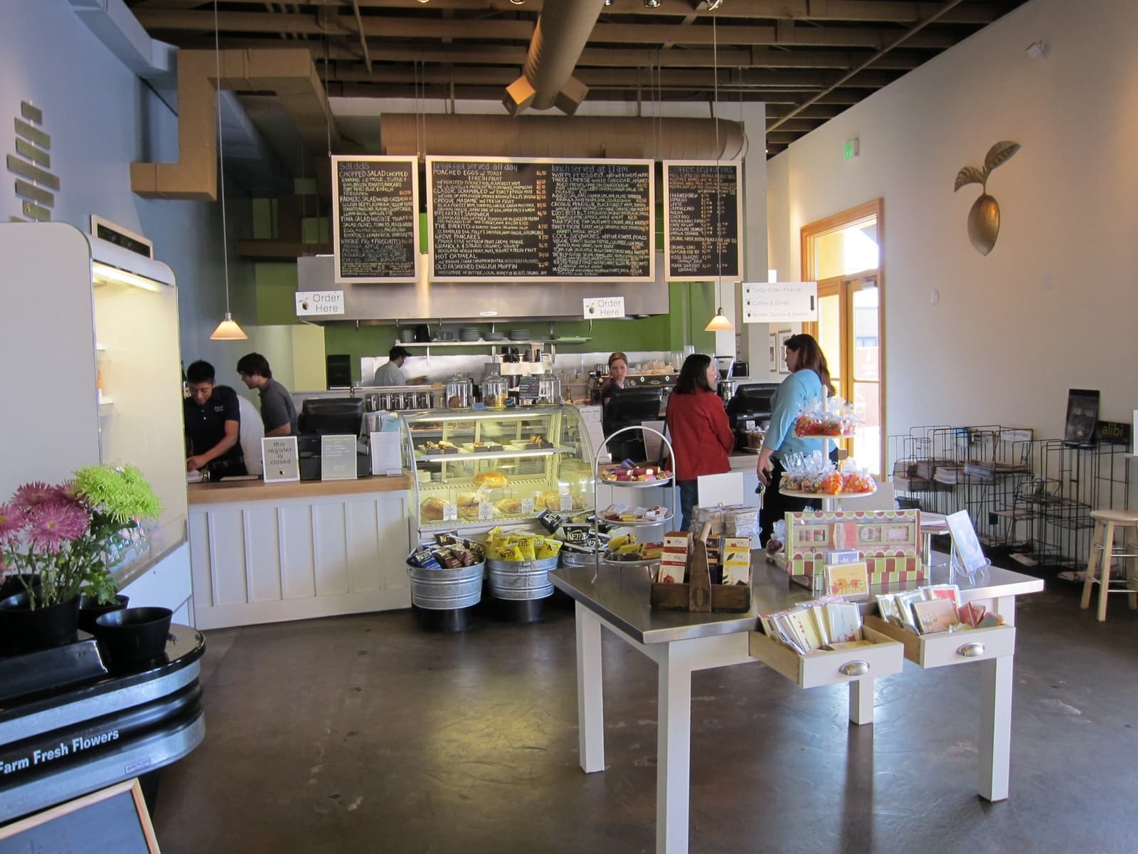 A café counter under chalkboard menus, with a market table in front