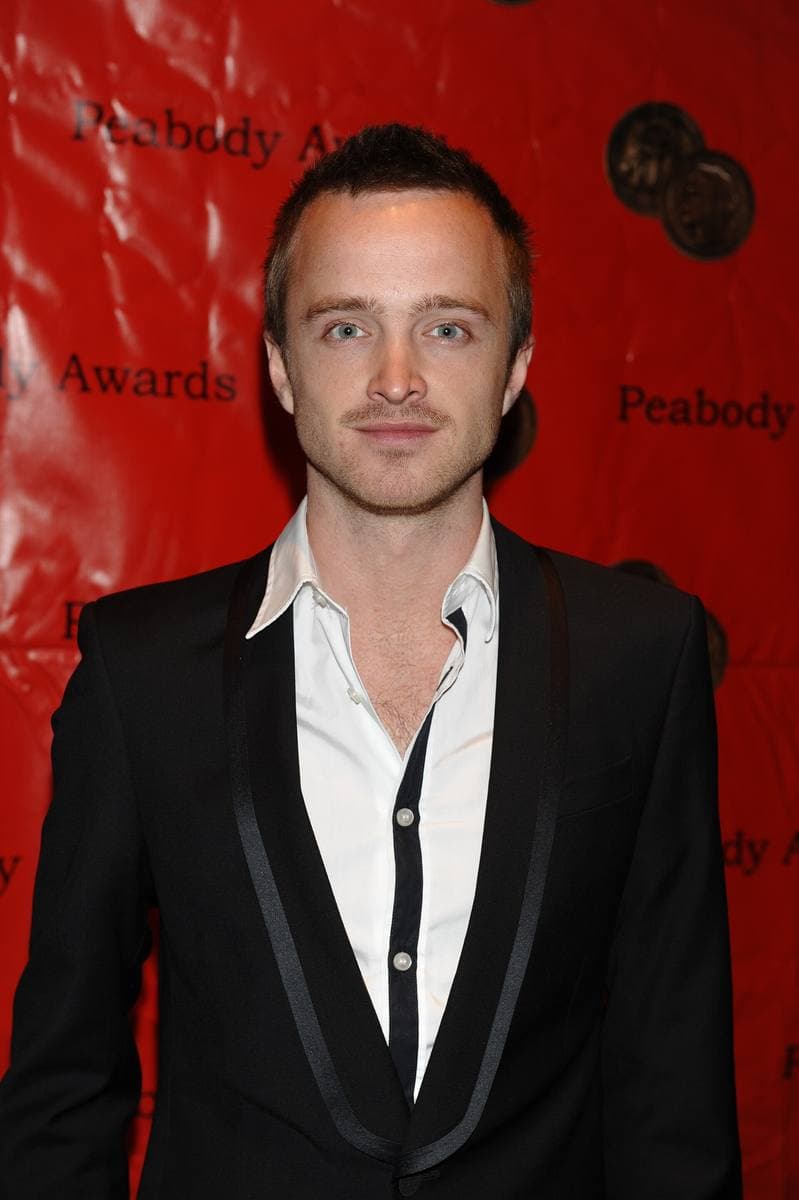 Aaron Paul in a black suit and open-necked shirt against a red backdrop