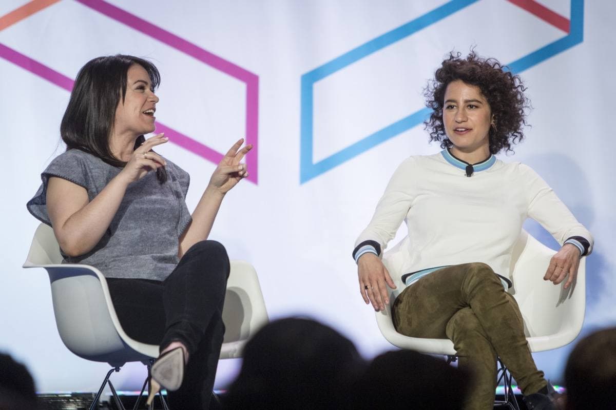 Two women in white chairs on a stage: one with long dark hair gesturing as she talks, one with curly hair laughing