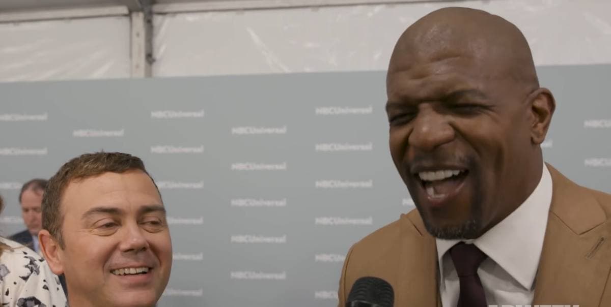 Two men in suits laughing in front of a pale branded backdrop