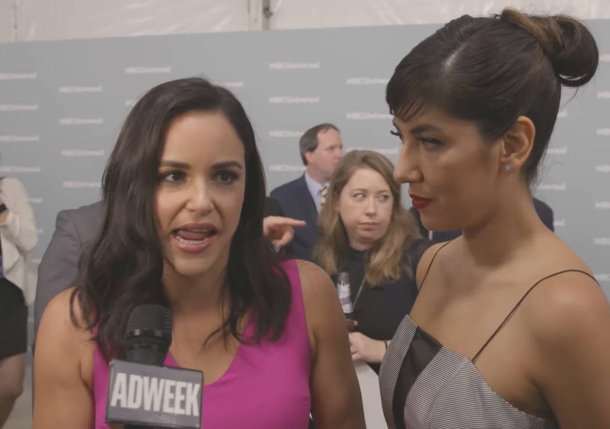 Two women talking to an interviewer in front of a pale branded backdrop, one laughing mid-sentence