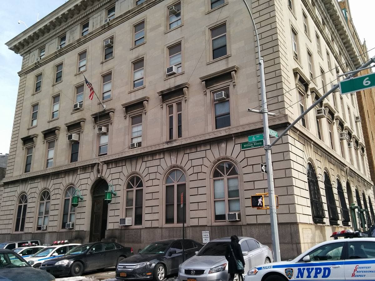A pale stone and brick police station house on a Brooklyn corner, with arched ground-floor windows and bare winter trees