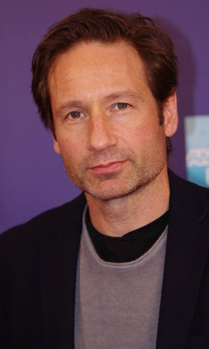 David Duchovny in a dark jacket and open shirt, smiling, against a dark backdrop