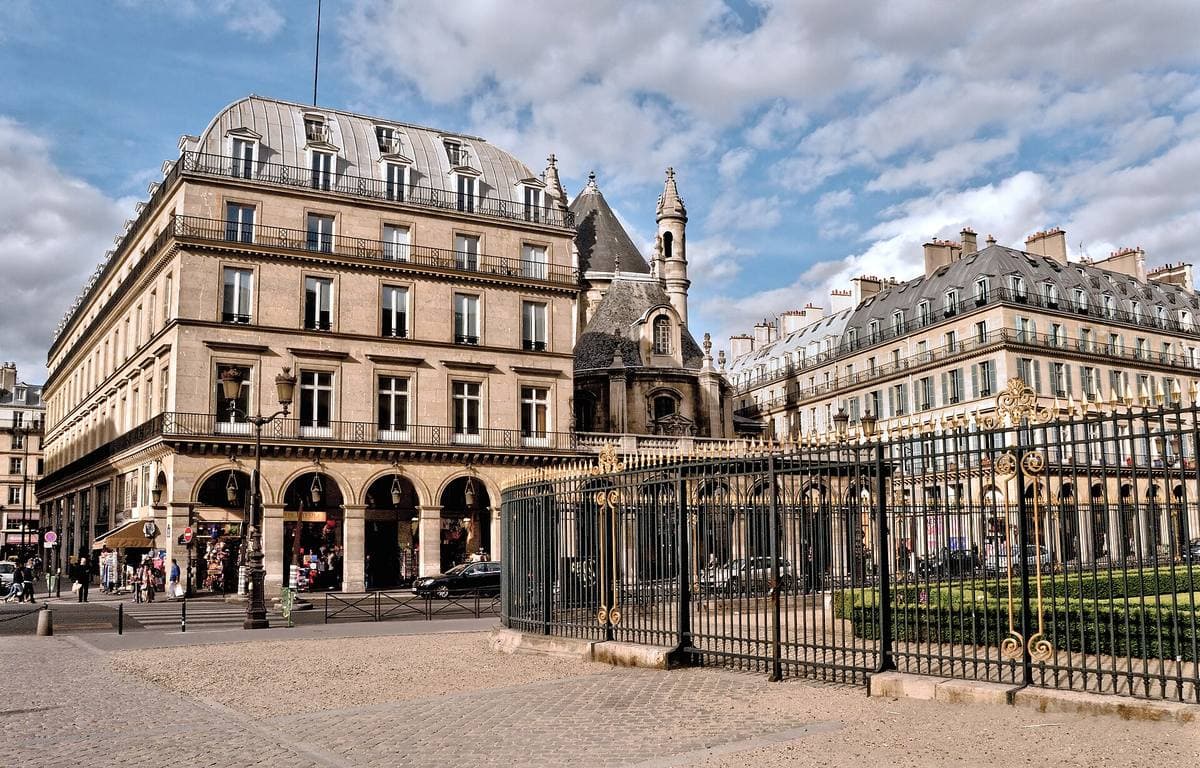 An arcaded stone building on a Paris corner under a blue sky, a church spire behind