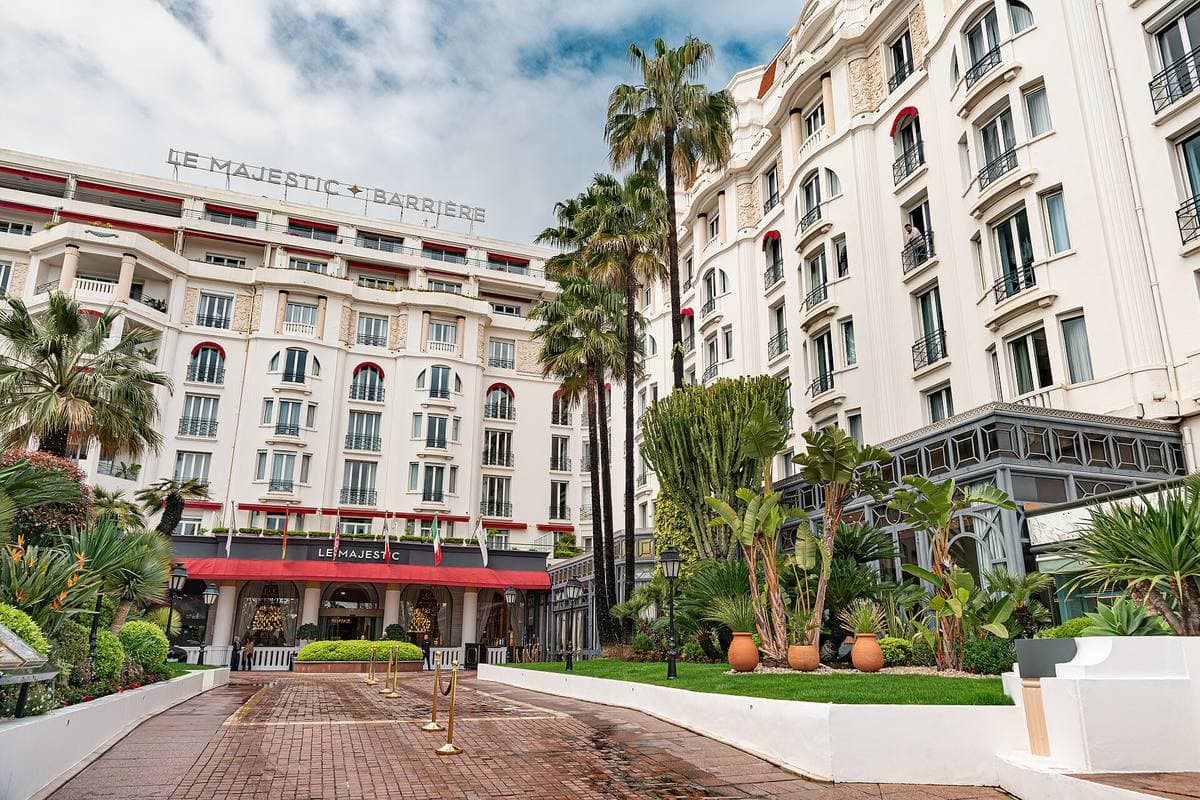 A white Riviera palace hotel behind palm trees and a red awning