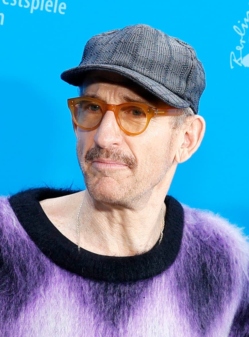 Johan Renck in a cap and round glasses against a blue backdrop