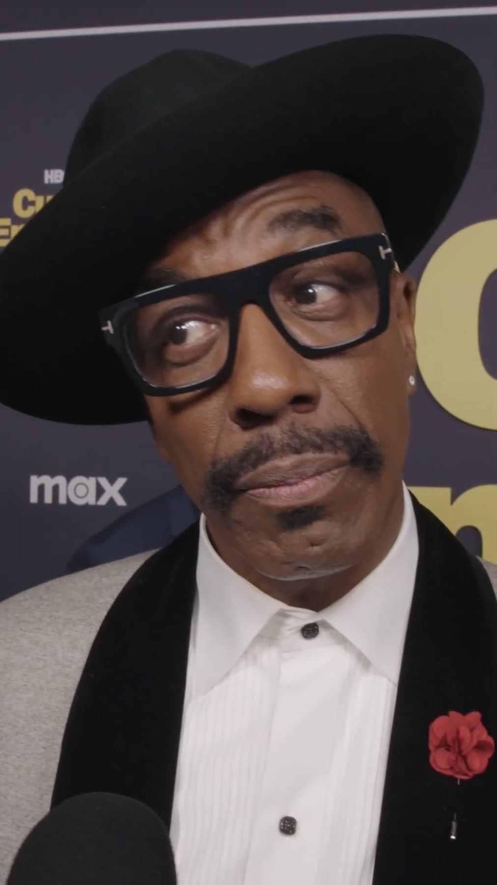 J. B. Smoove in a black hat and thick-framed glasses, eyebrow raised