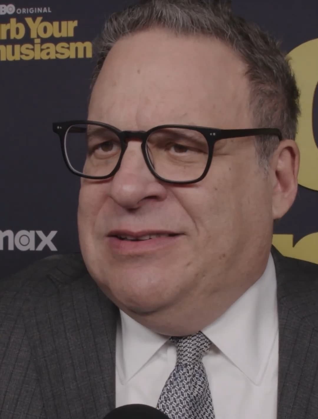 Jeff Garlin in black glasses, a grey suit and a patterned tie