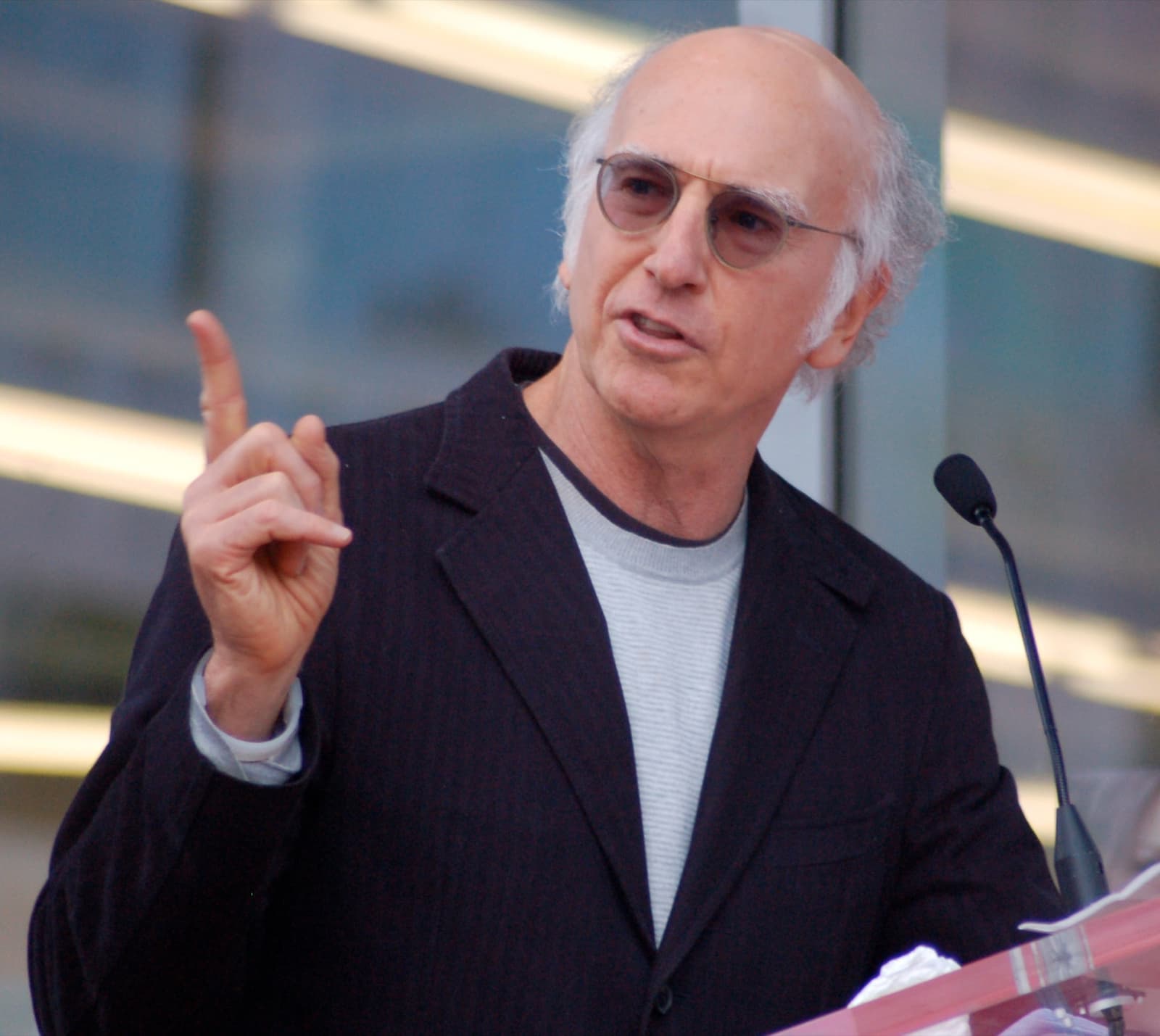 Larry David at a microphone, one finger raised as if making a point