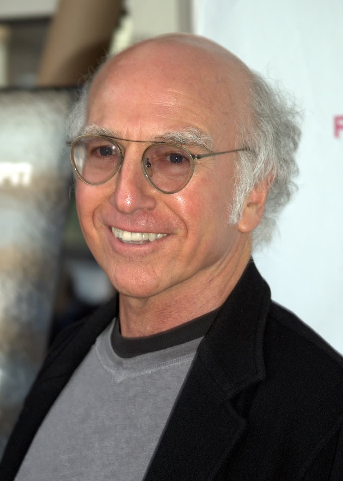 Larry David smiling in round glasses, a grey T-shirt and a dark jacket