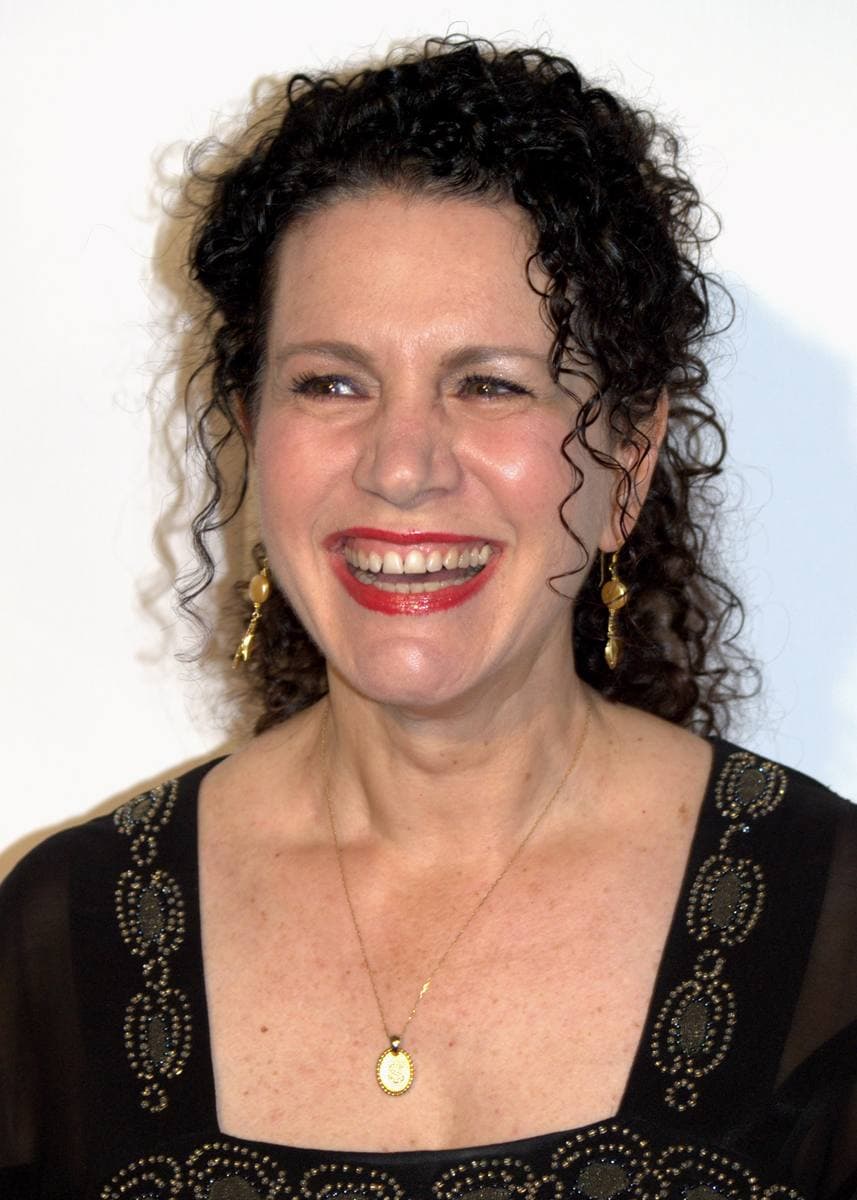 Susie Essman laughing, dark curly hair, red lipstick and a black beaded top, against a white wall.