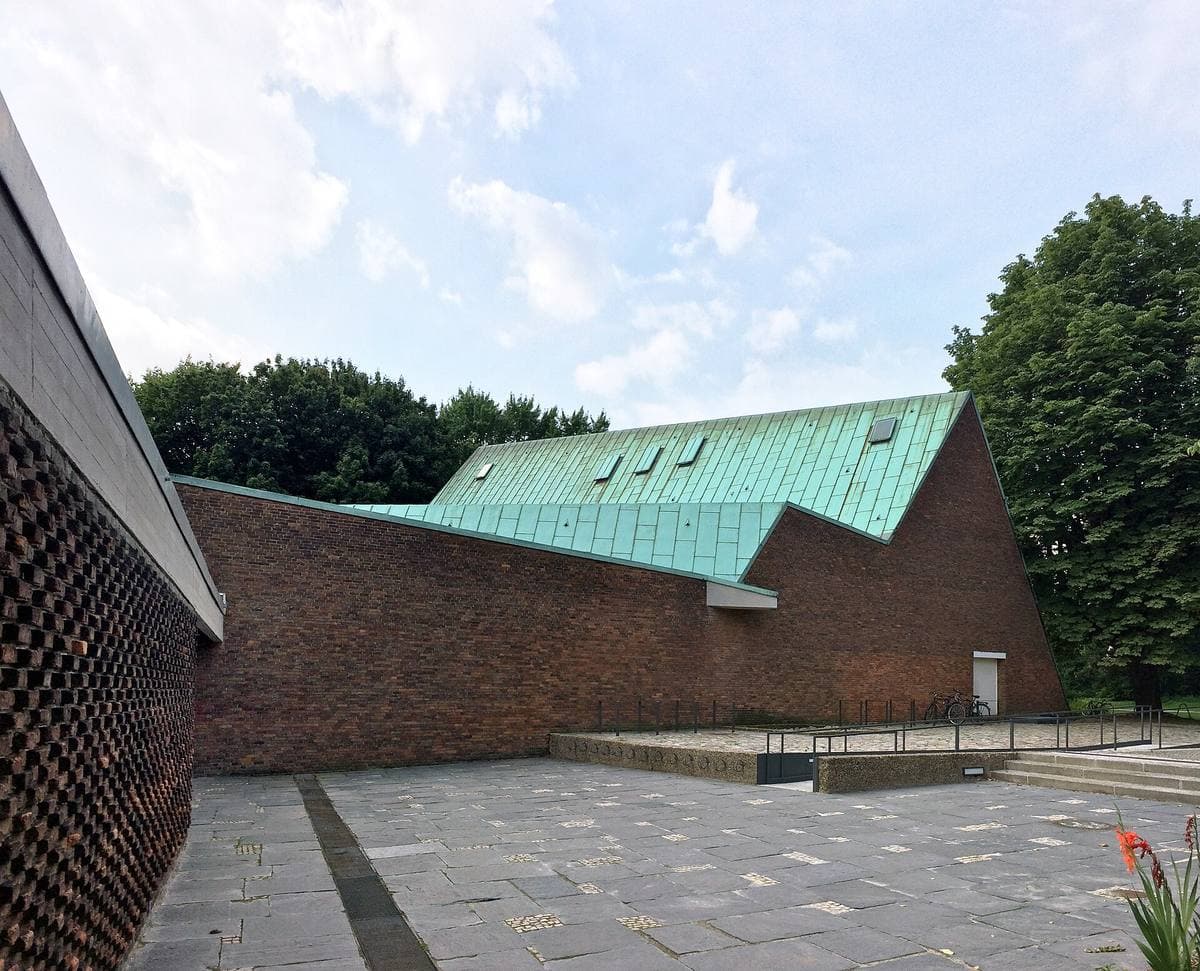 A low brick building with a green copper roof behind a paved forecourt