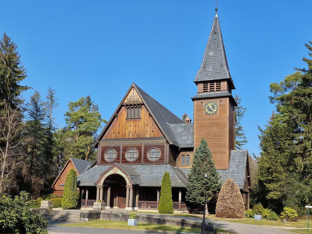 A dark timber chapel with a steep slate roof and a clock tower among pines under a blue sky
